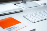 A white desk holds a keyboard, a closed laptop, a computer monitor, a wireless mouse, and an insightful book titled "Brand Identity" with its striking orange and white cover.