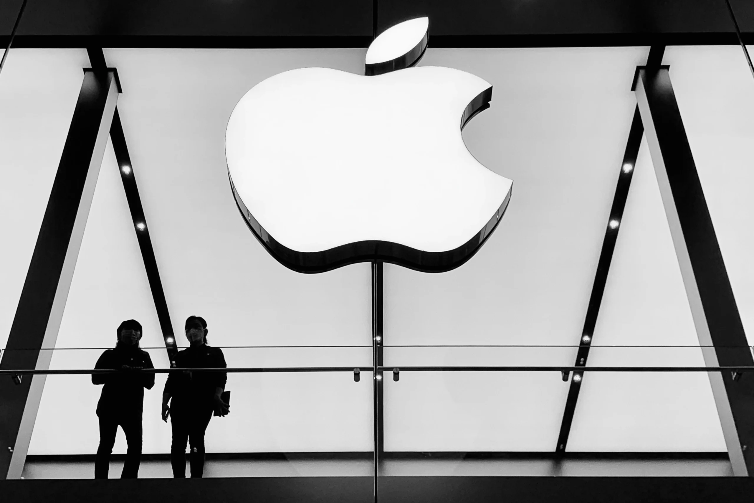 Two people stand on a glass balcony beneath a large illuminated Apple logo in a monochrome setting, perfectly capturing the brand identity with its sleek and minimalist design.